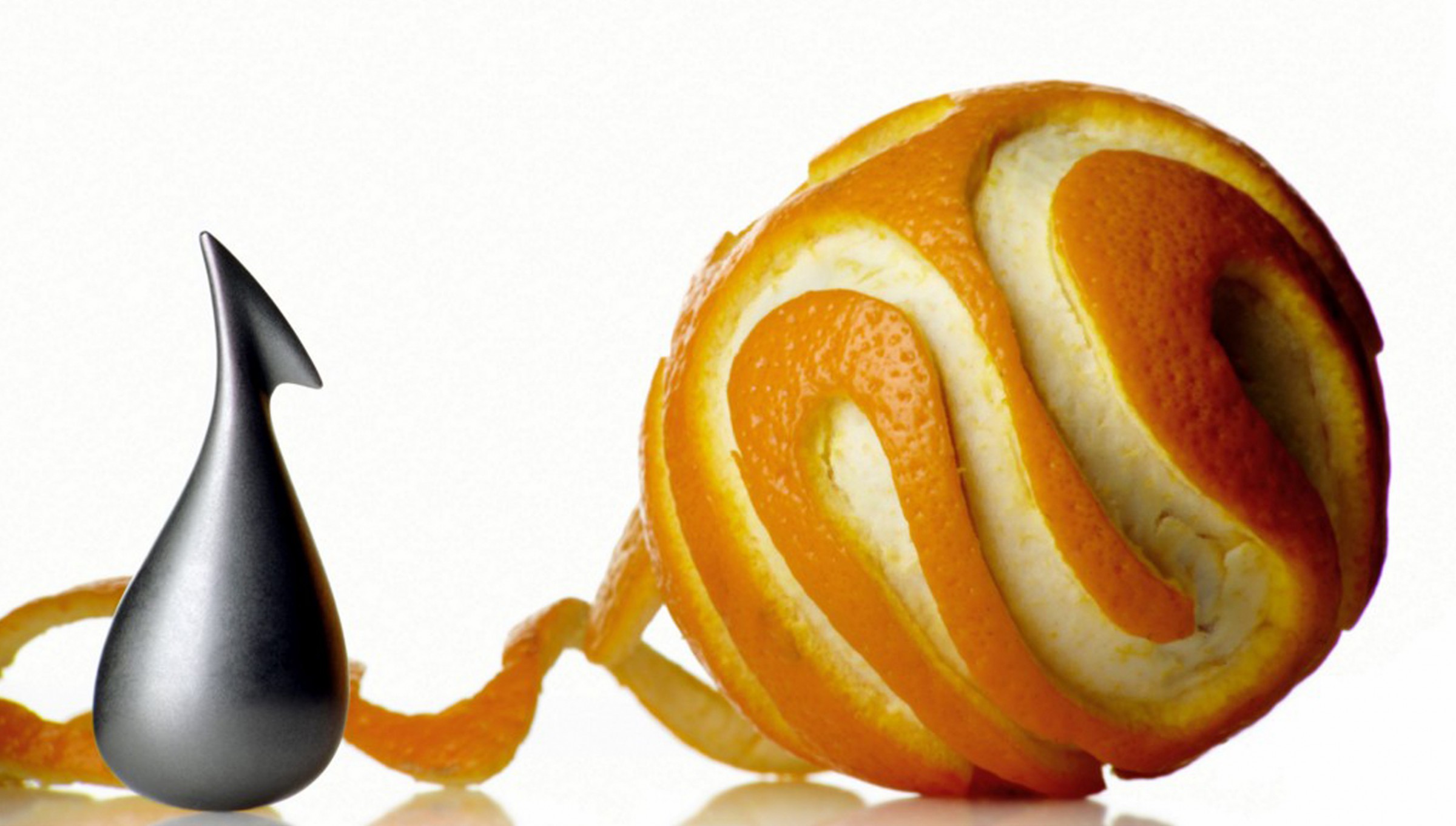 A grey Alessi Apostrophe Orange Peeler stands beside an artfully peeled orange